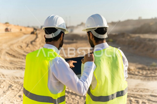 تصوير من الخلف لمقاول ومشرف العمل يرتديان خوذة وسترة خاصة بالعمل، عربيان خليجيان سعوديان يستخدمان الحاسوب المحمول في موقع العمل ، مناقشة العمل ،اشراف ومتابعة العمل ، متابعة استراتيجية المشروع ، التصاميم المعمارية والانشائية