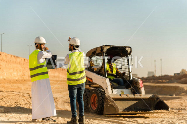 تصوير من الخلف لمقاول ومشرف العمل يرتديان خوذة وسترة خاصة بالعمل، عربيان خليجيان سعوديان يستخدمان الحاسوب المحمول في موقع العمل ، مناقشة العمل ،اشراف ومتابعة العمل ، متابعة استراتيجية المشروع ، التصاميم المعمارية والانشائية