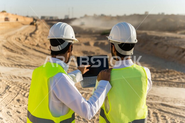 تصوير من الخلف لمقاول ومشرف العمل يرتديان خوذة وسترة خاصة بالعمل، عربيان خليجيان سعوديان يستخدمان الحاسوب المحمول في موقع العمل ، مناقشة العمل ،اشراف ومتابعة العمل ، متابعة استراتيجية المشروع ، التصاميم المعمارية والانشائية