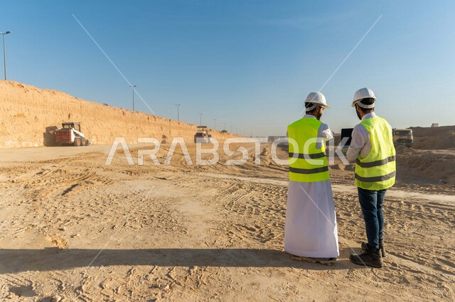 تصوير من الخلف لمقاول ومشرف العمل يرتديان خوذة وسترة خاصة بالعمل، عربيان خليجيان سعوديان يستخدمان الحاسوب المحمول في موقع العمل ، مناقشة العمل ،اشراف ومتابعة العمل ، متابعة استراتيجية المشروع ، التصاميم المعمارية والانشائية
