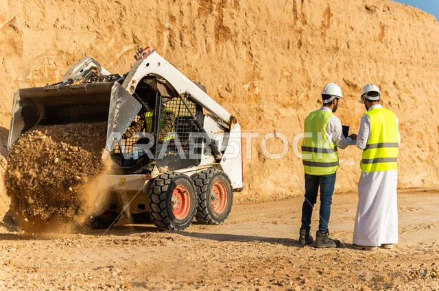تصوير من الخلف لمقاول ومشرف العمل يرتديان خوذة وسترة خاصة بالعمل، عربيان خليجيان سعوديان يستخدمان الحاسوب المحمول في موقع العمل ، مناقشة العمل ،اشراف ومتابعة العمل ، متابعة استراتيجية المشروع ، التصاميم المعمارية والانشائية