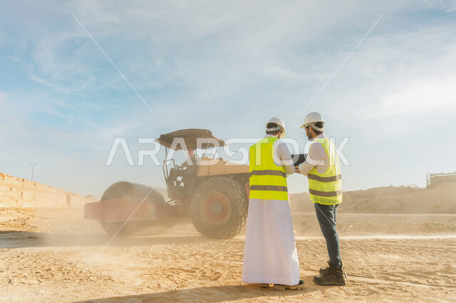 تصوير من الخلف لمقاول ومشرف العمل يرتديان خوذة وسترة خاصة بالعمل، عربيان خليجيان سعوديان يستخدمان الحاسوب المحمول في موقع العمل ، مناقشة العمل ،اشراف ومتابعة العمل ، متابعة استراتيجية المشروع ، التصاميم المعمارية والانشائية