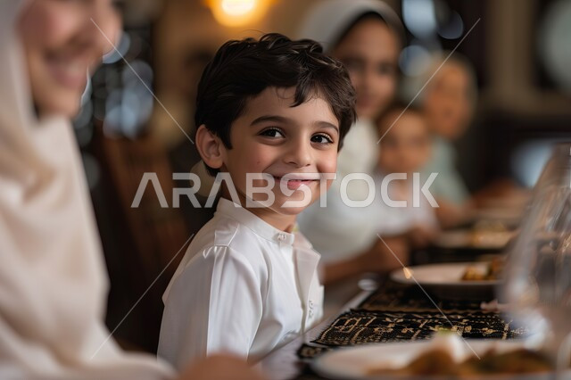 قضاء أوقات أسرية ممتعة وقت العطلة، صورة مقربة لولد عربي سعودي خليجي يجلس على مائدة الطعام في أحد المطعم الراقية، النظر الى الكاميرا بايماءات السعادة، انتظار وجبة الطعام بحماس، أجواء عائلية ترفيهية، مفهوم المحبة والمودة بين أفراد الأسرة 