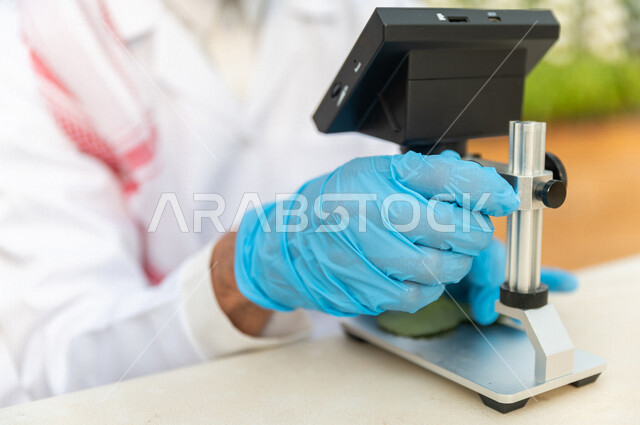 مهندس زراعي عربي خليجي سعودي ، يرتدي الروب الطبي والقفازات الطبية ،يستخدم جهاز المايكروسكوب لفحص الاوراق النباتية ، عينة من الاوراق النباتية ، الرقابة على المشاتل الزراعية