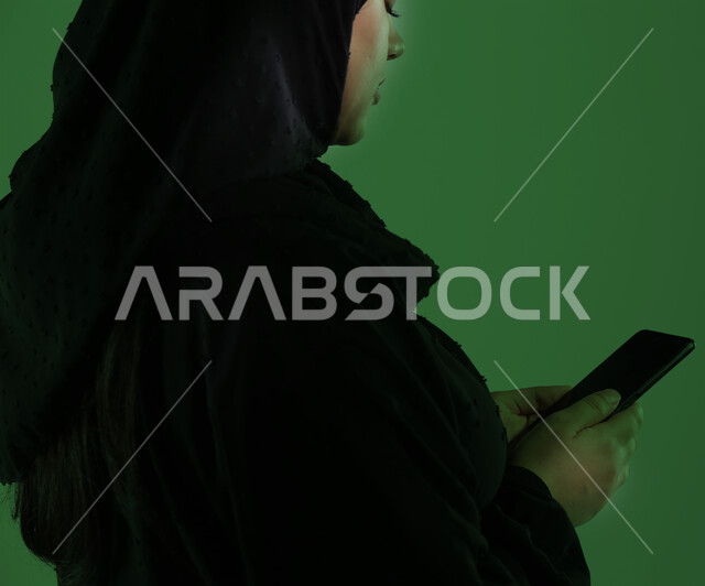 Using modern technology devices for entertainment and fun, browsing social media via mobile, veiled Saudi Arabian Gulf woman wearing black abaya holding mobile phone in hand, green chroma key background