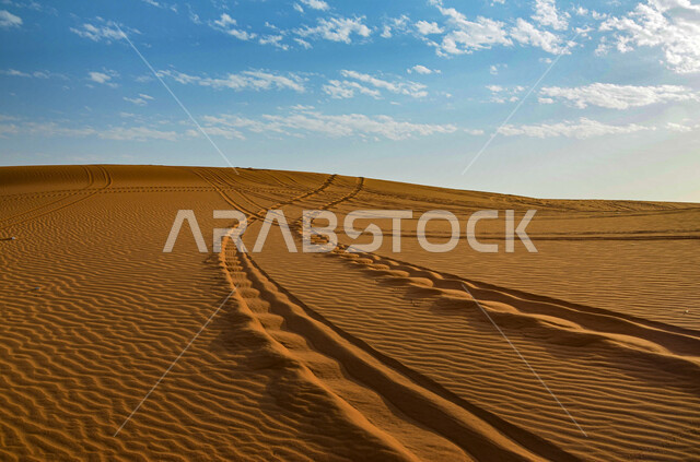 البيئة الطبيعية الخليجية الجافة، تشكل الكثبان والقمم الرملية في الصحاري, المساحات الصحراوية الشاسعة الممتدة على طول الأفق, مشهد لتداخل ألوان رمال الصحراء الذهبية اللامعة مع خلفية السماء الزرقاء الملبدة بالغيوم 