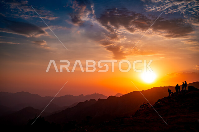 القيام بمغامرات ورحلات السفاري, صورة ظلية لمجموعة من الأشخاص العربي الخليجيين السعوديين يقفون على قمة جبل الصفا في مدينة الطائف, مراقبة غروب الشمس من المرتفعات الجبلية والاستمتاع بالمناظر الطبيعية, خلفية السماء البرتقالية الملبدة بالغيوم