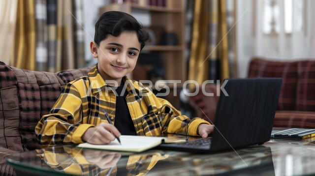 الاجتهاد والمشاركة التطوعية من خلال دورات التعلم عن بعد، بورتريه لتلميذ عربي خليجي سعودي, يحضر دروسه عن طريق الكمبيوتر، طالب  يجلس على طاولة الدارسة ويمسك بيده القلم ويضع الدفتر امامه