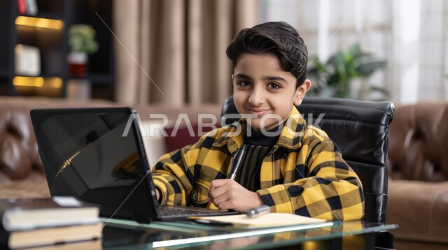 طالب يجلس على طاولة الدارسة ويمسك بيده القلم ويضع الدفتر امامه, الاجتهاد والمشاركة التطوعية من خلال دورات التعلم عن بعد، بورتريه لتلميذ عربي خليجي سعودي, يحضر دروسه عن طريق الكمبيوتر