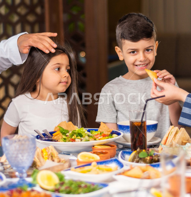 صورة مقربة لطفلان عربيان خليجيان سعوديان مبتسمان وسعيدان ، يتناولان الطعام في المطعم ، مائدة طعام مليئة بأشهى المأكولات والمشروبات ، قضاء قت ممتع ، اجواء عائلية سعيدة