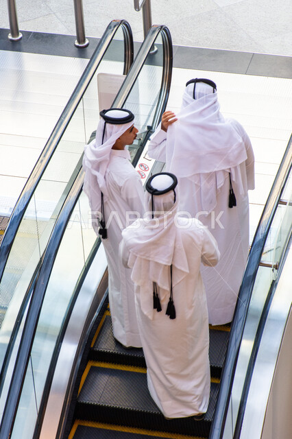 التصاميم والديكورات الداخلية المتميزة داخل دبي مول، مراكز التسوق والمتاجر الفاخرة، صورة من الاعلى لمجموعة رجال عرب خليجيي اماراتيين يرتدون القندورة والغترة يصعدون على الدرج الكهربائي، السياحة بالإمارات العربية المتحدة، اماكن الأنشطة العائلية الترفيهية