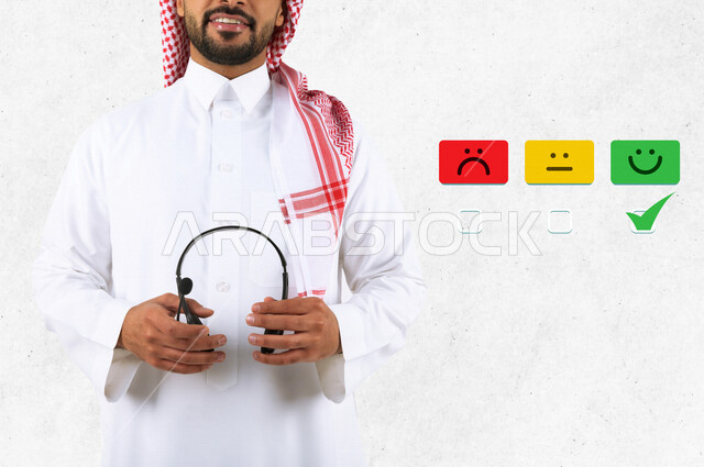 صورة مقربة لرجل عربي خليجي سعودي مبتسم ، موظف خدمة العملاء يمسك بيده سماعات ، يقف بجوار مجموعة من  أيقونات تقييم الاداء ( أداء جيد ، أداء متوسط ، أداء سئ ) ، كسب رضا العملاء .