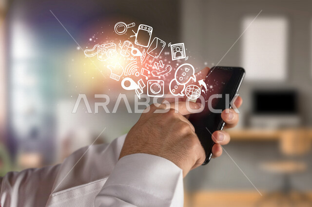 صورة مقربة لرجل عربي خليجي سعودي يمسك جوال حديث بيده و يضغط بأصبعه ، خروج مجموعة من الأيقونات الخاصة بتطبيقات الجوال و الأجهزة الالكترونية المتطايرة بتقنية الهولوجرام ، اسستخدام تقنية الهولوجرام ثلاثية الأبعاد .