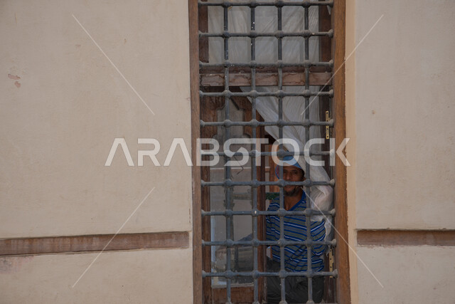 الفن الهندسي المعماري على الطراز العربي القديم، أماكن سياحية تراثية شعبية، رجل عربي خليجي سعودي يقف خلف نافذة خشبية مغلقة بشبك حديدي، المدينة التاريخية في المملكة العربية السعودية، االمباني والبيوت الأثرية في البلد التاريخي في جدة, المعالم التاريخية في حي البلد