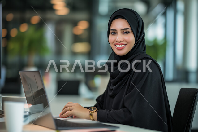 Comfortable work environment in Saudi companies, women's office jobs and professions, looking at the camera with gestures of pleasure and happiness to accomplish tasks, business management and organization, smiling veiled Saudi Arabian Gulf woman using a laptop