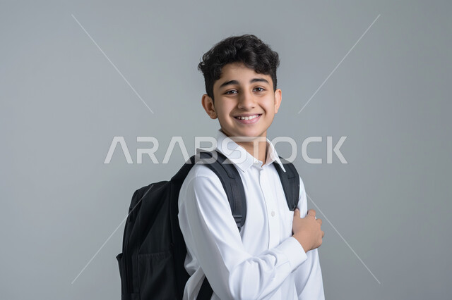 العوده إلى المدارس, النظر الى الكاميرا بايماءات السعادة والسرور، تعابير الأدب والأخلاق, حب التعلم والتعليم, بورتريه مقرب لطالب عربي خليجي سعودي يرتدي الثوب التقليدي ويحمل حقيبة الظهر المدرسية, الإستعداد للعام الدراسي الجديد, خلفية رمادية
