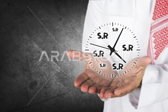 صورة مقربة لرجل عربي خليجي يحمل بين يديه  شكل ساعة تحتوي على رمز اختصار الريال السعودي ، تتبع البورصة المالية صعودها و هبوطها .