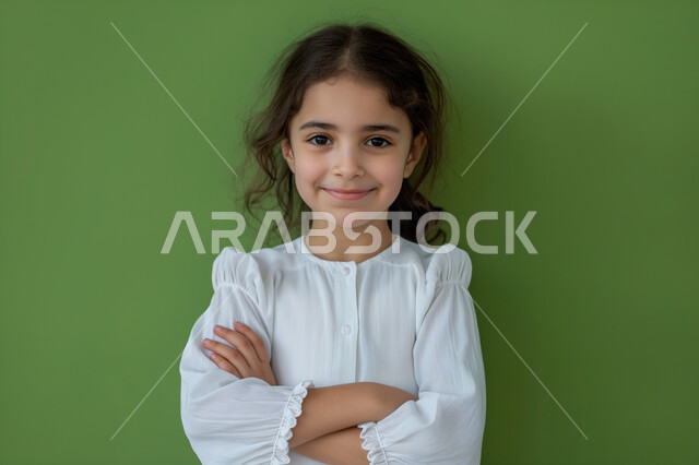 ملامح طفولية بريئة، الأناقه والاهتمام بالمظهر الخارجي، بورتريه مقرب لفتاة عربية سعودية خليجية مبتسمة ترتدي فستان أبيض تنظر الى الكاميرا بايماءات السعادة والبشاشة، الوقوف باستقامة بأيدي متقاطعة بتعابير الثقة بالنفس، التقاط صور شخصية للوثائق الرسمية، خلفية خضراء