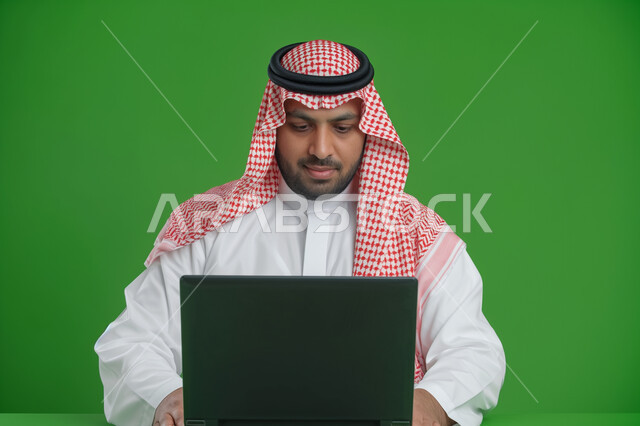 إجراء مقابلات عمل أونلاين, استخدام الأجهزة والتقنيات الحديثة لتسهيل الأعمال, مهن ووظائف سعودية مكتبية، ايماءات وتعابير التركيز والاندماج، بورتريه مقرب لرجل أعمال عربي سعودي خليجي يرتدي الزي التقليدي والشماغ يجلس على مكتب أمام  جهاز الحاسوب المحمول, خلفية خضراء