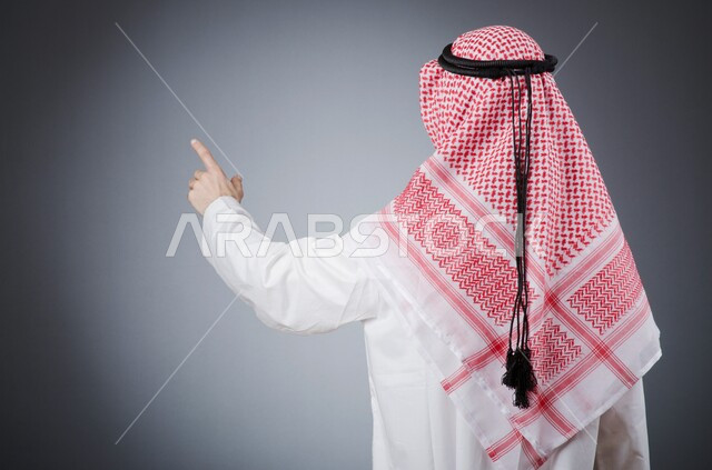 Pointing to something with gestures of identification and emphasis, portrait from behind of a Saudi Arabian Gulf man wearing traditional dress and shemagh raising index finger with expressions of touching an item, gray background