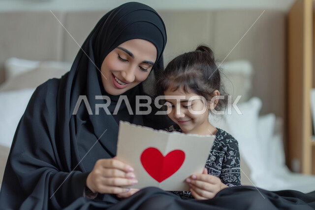 Presenting greeting cards and gifts for holidays and occasions, commemorating Mother's Day, the concept of respecting and appreciating parents, a veiled Saudi Arabian Gulf woman wearing the traditional abaya holding a greeting card given to her by her little girl, a mother embracing her daughter in an atmosphere full of love, affection and tenderness