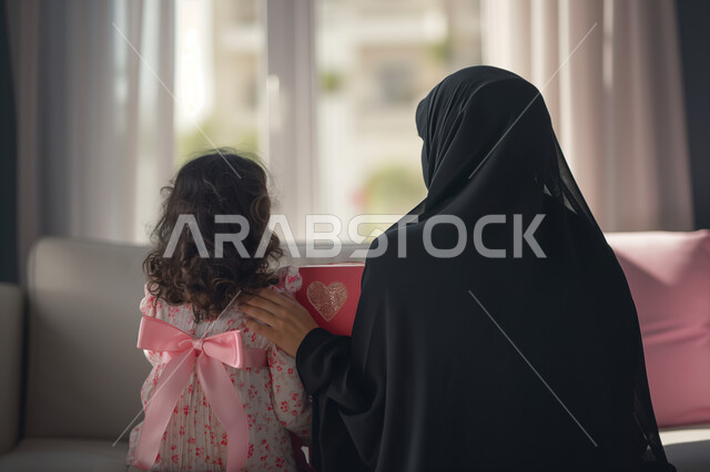  التهنئة بالأعياد والمناسبات السعيدة ، إحياء ذكرى عيد الأم، مفهوم احترام وتقدير الوالدين، صورة من الخلف لامرأة عربية خليجية سعودية محجبة ترتدي العباءة التقليدية تجلس بجانب طفلتها الصغيرة بجو من الحب والحنان، تقديم الهدايا والمعايدات والمباركات من قبل الأبناء