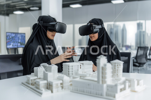 Using virtual reality to enhance the idea of ​​architectural planning, using modern electronic technologies to improve work efficiency, a close-up image of two Saudi Arabian Gulf women wearing hijab and abayas and wearing VR glasses, explanation of miniature models of towers and skyscrapers