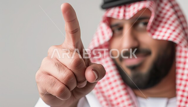 تعابير التحديد والتأكيد, الاشارة إلى شيء ما بإيماءات السرور ,بورتريه لرجل عربي خليجي سعودي مبتسم يرتدي الزي التقليدي والشماغ يشير بأصابع السبابة للاختيار وينظر إلى الكاميرا، خلفية رمادية