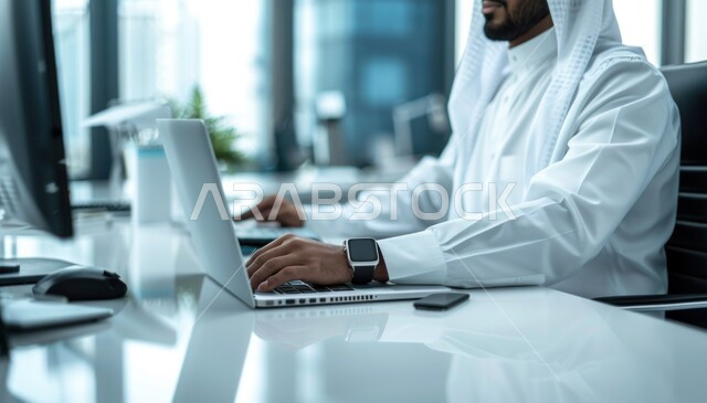 إدارة شؤون الأعمال وتنظيمها عبر الاجهزة التقنية، مهن ووظائف ادارية مكتبية، دمج العمل مع التقنية والتكنولوجيا، رجل اعمال عربي خليجي سعودي يرتدي الزي التقليدي يعمل على الحاسوب المحمول داخل المكتب