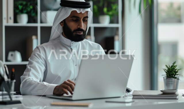 إدارة شؤون الأعمال وتنظيمها عبر الاجهزة التقنية، مهن ووظائف ادارية مكتبية، دمج العمل مع التقنية والتكنولوجيا، رجل اعمال عربي خليجي سعودي يرتدي الزي التقليدي يعمل على الحاسوب المحمول داخل المكتب