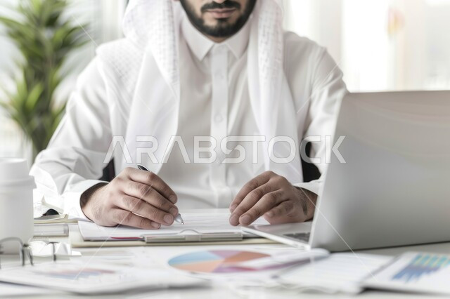 إدارة شؤون الأعمال وتنظيمها عبر الاجهزة التقنية، مهن ووظائف ادارية مكتبية، دمج العمل مع التقنية والتكنولوجيا، رجل اعمال عربي خليجي سعودي يرتدي الزي التقليدي يعمل على الحاسوب المحمول داخل المكتب
