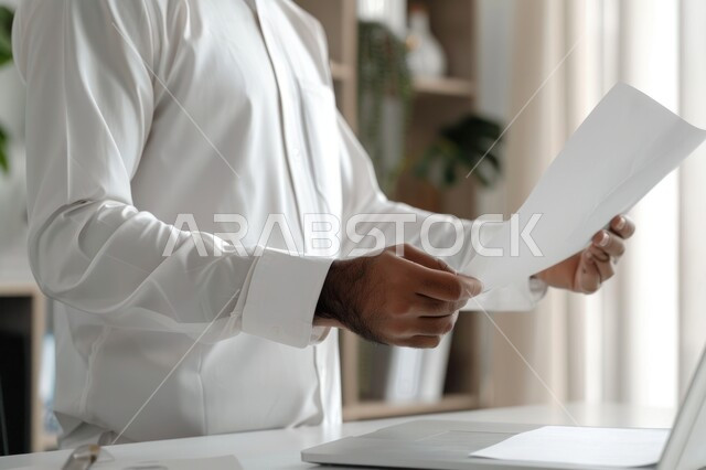 استخدام الأجهزة والتقنيات الحديثة لإنجاز المهام، مهن وأعمال مكتبية تجارية، رجل أعمال سعودي خليجي يرتدي الزي التقليدي يعمل على جهاز الحاسوب المحمول، مفهوم إدارة الأعمال، عقد صفقات عمل عبر الإنترنت، مراجعة ومتابعة البحوث العلمية وخطة العمل في مقر الشركة