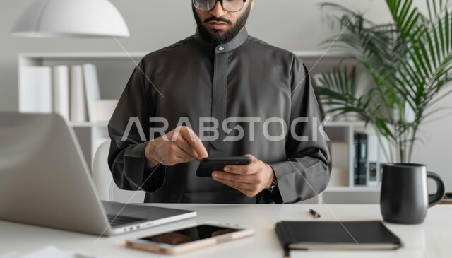 مهن ووظائف ادارية مكتبية, إنجاز المهام العمل المطلوبة ، دمج العمل مع التقنيات والتكنولوجيا، مفهوم إدارة شؤون الأعمال وتنظيمها عبر الاجهزة التقنية، رجل اعمال عربي خليجي سعودي يرتدي زي تقليدي أسود يمسك الهاتف المحمول في المكتب