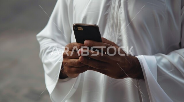 البحث عن عمل عن طريق الانترنت، تصفح مواقع التواصل الإجتماعي، بورتريه مقربة لرجل عربي خليجي سعودي يرتدي الزي التقليدي يحمل بيده جهاز الهاتف المحمول، التسويق الإلكتروني عبر الأجهزة الحديثة، متابعه آخر أخبار السوشال ميديا، خلفية رمادية