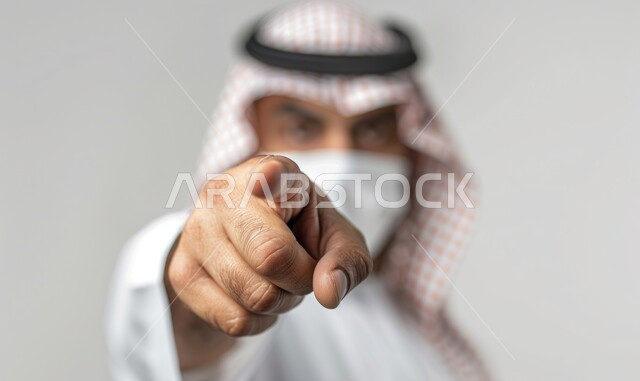  الاشارة إلى شيء ما, النظر الكاميرا بتعابير العصبية والغضب، بورتريه لرجل عربي خليجي سعودي يرتدي الزي التقليدي والشماغ ويضع كمامة يشير بأصبع السبابة لتوجيه الاتهام، ايماءات التحذير والتنبيه، خلفية بيضاء