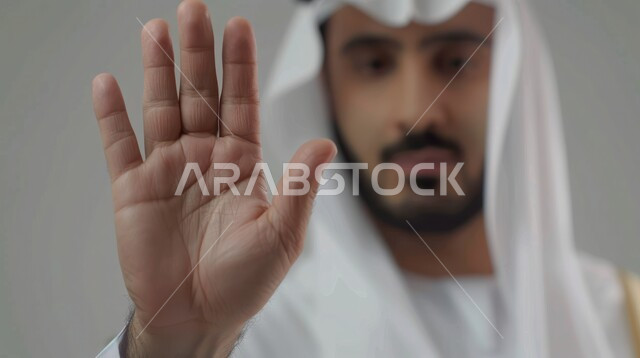 طلب عدم التدخل وخفض الصوت والتهديد, الوقوف بغضب ورفض أمر محدد, الصمت والتوقف عن الكلام, الامتناع عن فعل أمر معين, بوتريه مقرب لرجل عربي خليجي سعودي يرتدي الزي التقليدي يرفع كفه للأعلى بإيماءات الانزعاج, خلفية رمادية