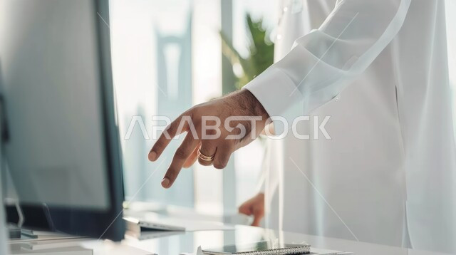 Using modern technologies and advanced devices, completing tasks via computer, developing and following up on administrative work, working in the office field, a Saudi Arabian Gulf employee wearing traditional clothing points to the computer screen