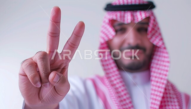 الفوز والنصر والحب, علامات وإشارات مشعة بالأمل والتفاؤل, الوقوف باستقامة والنظر إلى الكاميرا, بورتريه لشاب عربي خليجي سعودي يرتدي الثوب التقليدي يرفع يده بعلامة ثنائية, المثابرة والتميز والتحديات لتحقيق النجاح, خلفية رمادية