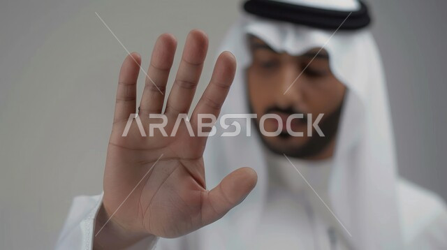 الوقوف بانزعاج ورفض أمر محدد, الصمت والتوقف عن الكلام, الامتناع عن فعل أمر معين, بورتريه مقرب لرجل عربي خليجي سعودي يرتدي الغترة والعقال يرفع كفه للأعلى بإيماءات طلب عدم التدخل وخفض الصوت والتهديد, خلفية رمادية