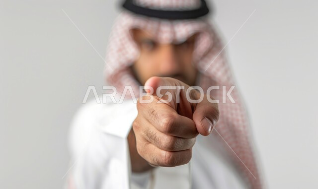 التوعد لشيء ما، النظر إلى الكاميرا بإيماءات الغضب, بورتريه مقرب لرجل عربي خليجي سعودي يرتدي الثوب التقليدي والشماغ يشير باصبع السبابة للأمام بالاتجاه المقابل, التهديد والمواجهة والمنافسة, خلفية رمادية