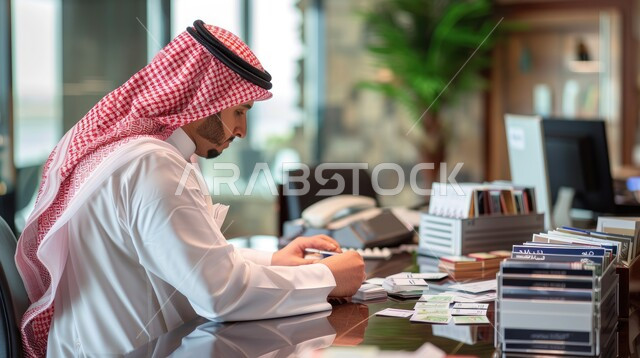 The concept of focusing on work, completing tasks and achieving goals, taking and reviewing notes, business files and data, a back view of a Saudi Arabian Gulf man wearing a traditional shemagh and thobe reviewing documents sitting in his office