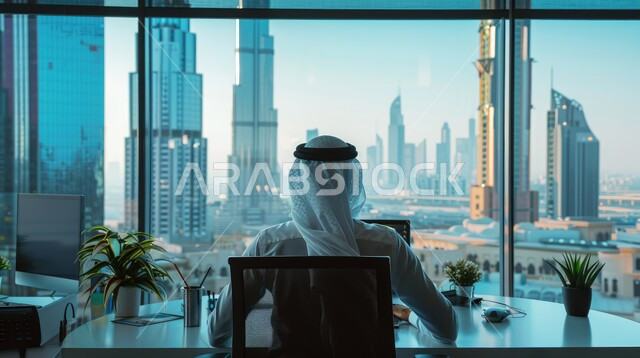 مفهوم التركيز على العمل, انجاز المهام وتحقيق الاهداف, استخدام التكنولوجيا والتقنيات الحديثة, صورة من الخلف لرجل عربي خليجي سعودي يرتدي العقال والثوب التقليدي يعمل على جهاز الحاسب المحمول يجلس داخل مكتبه، خلفية أبراج المملكة