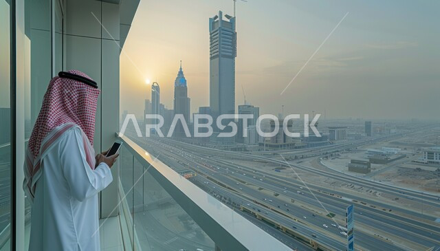 التطور والتقدم العمراني السعودي, معالم وأماكن سياحية مشهورة, إطلالة على الأبراج وناطحات السحاب، الاستمتاع بالمناظر الطبيعية, مناطق جذب الزوار، صورة من الخلف لرجل عربي خليجي سعودي يرتدي الشماخ ويحمل هاتف محمول يقف على الشرفة