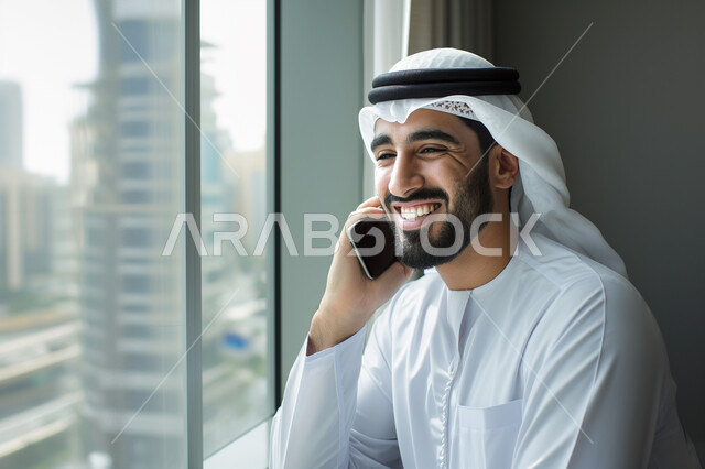 التواصل مع الأهل والأصدقاء، إيماءات وتعابير السعادة والسرور لسماع الأخبار المفرحة، صورة مقربة لرجل عربي خليجي إماراتي مبتسم يرتدي الكندورة والغترة يقوم بإجراء مكالمة هاتفية عبر الهاتف المحمول، استخدام التقنيات الحديثة باختصار المسافات وإزالة الشوق بين الأقارب 