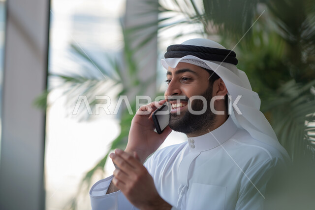 التواصل مع الأهل والأصدقاء، إيماءات وتعابير السعادة والسرور لسماع الأخبار المفرحة، صورة مقربة لرجل عربي خليجي إماراتي مبتسم يرتدي الكندورة والغترة يقوم بإجراء مكالمة هاتفية عبر الهاتف المحمول، استخدام التقنيات الحديثة باختصار المسافات وإزالة الشوق بين الأقارب 
