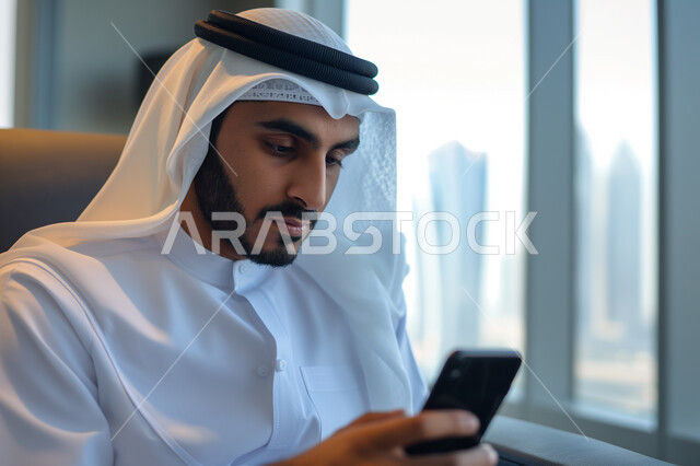 Integrating modern technologies and advanced devices into work, close-up of a smiling Saudi Arabian Gulf man wearing traditional thobe and shemagh sitting in his office using his mobile phone, chatting and communicating with family, friends and relatives