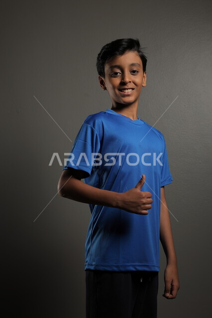  إيماءات و تعابير وجه و يدين تدل على الإعجاب و الرضى، بورتريه لطفل عربي خليجي سعودي يرتدي اللباس الرياضي باللون الازرق يرفع يده و يشير بأصبع الإبهام بإشارة الإعجاب، خلفية رمادي