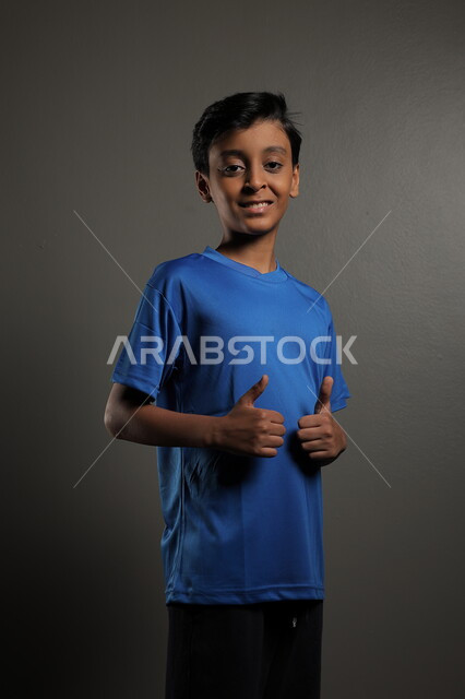  إيماءات و تعابير وجه و يدين تدل على الإعجاب و الرضى، بورتريه لطفل عربي خليجي سعودي يرتدي اللباس الرياضي باللون الازرق يرفع يده و يشير بأصبع الإبهام بإشارة الإعجاب، خلفية رمادي