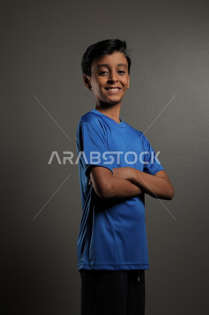 الوقوف بأيدي متقاطعة بتعابير الثقة بالنفس،  بورتريه لولد عربي خليجي سعودي يرتدي اللباس الرياضي باللون الازرق ينظر إلى الكاميرا، التقاط صور شخصية، الأناقه والاهتمام بالمظهر الخارجي، خلفية رمادية