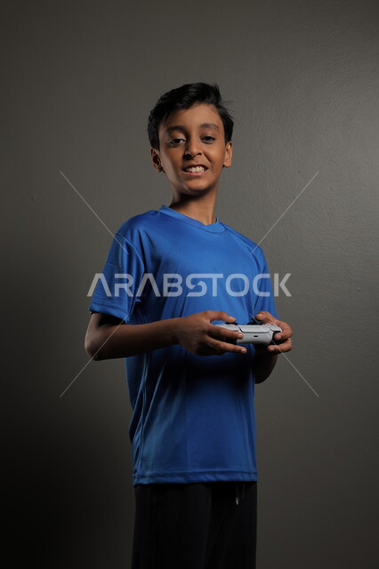 قضاء اوقات ممتعة باللعب في المنزل، استخدام التقنيات الحديثة للتسلية والترفيه، بورتريه لولد عربي خليجي سعودي يرتدي الزي الكاجوال يقوم باللعب في ألعاب الفيديو بايماءات الاندماج والتركيز، خلفية رمادية